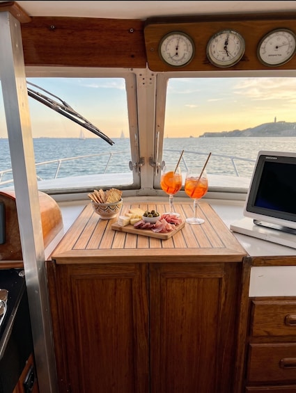 A boat's interior with a wooden table displaying food and drinks, a laptop, and a view of the sea and a distant island. 1