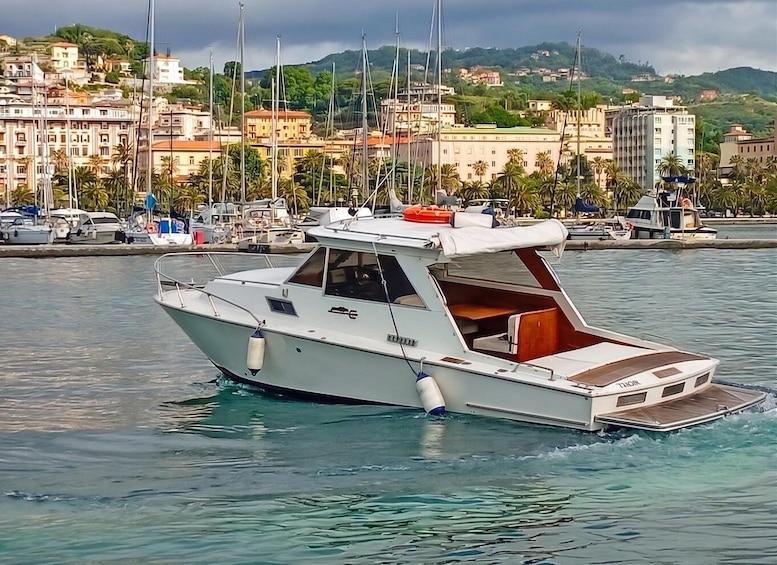 A white motorboat with a cabin and a red lifebuoy on the water, docked near a marina with buildings and hills in the background. 4