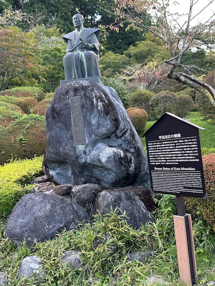A bronze statue of a seated figure on a rock, surrounded by greenery and a sign with text. 3