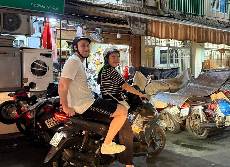 Two people on a motorcycle, a building with a sign, and a parked scooter. 3