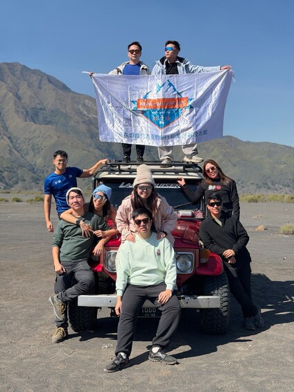 A group of people posing with a vehicle and a banner in a mountainous area. 2