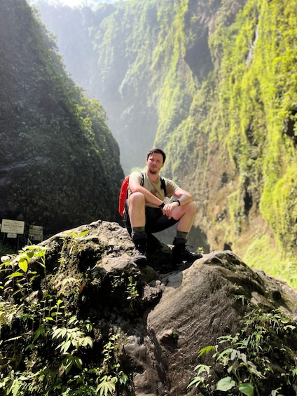 A person sitting on a rock with a red backpack, surrounded by lush greenery and steep cliffs. 4