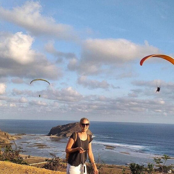 A person standing on a cliff overlooking the ocean with paragliders in the sky. 2