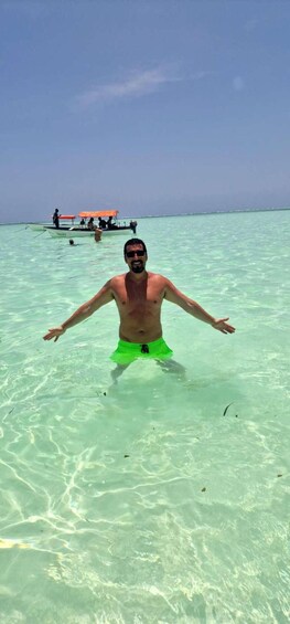A man in green swim trunks standing in shallow clear water with a boat and people in the background. 3