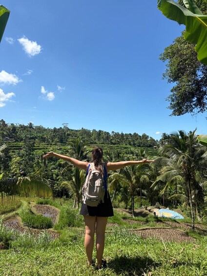 A person with arms outstretched in a lush, green landscape with palm trees and terraced fields. 5