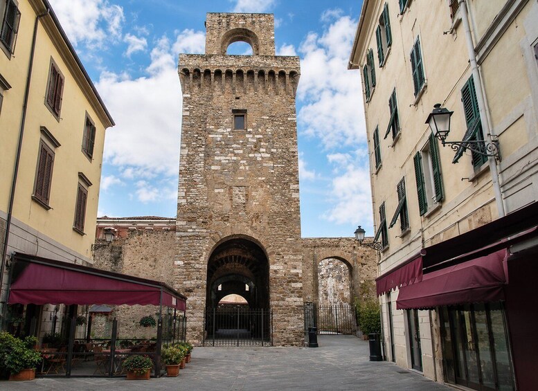 A stone tower with arched openings, flanked by buildings with shutters and outdoor seating. 3