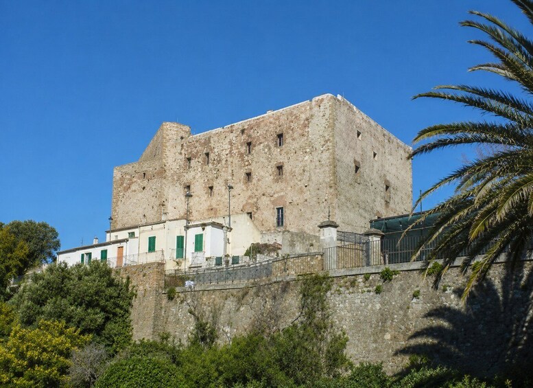A historic stone building with green shutters, surrounded by lush greenery and a clear blue sky. 2