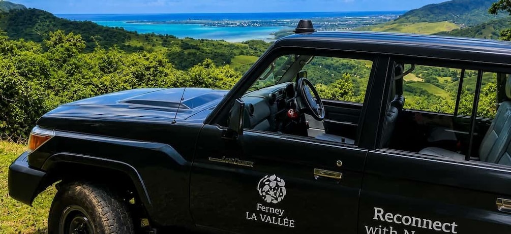 A black Land Rover with 'Ferney LA VALLEE' and 'Reconnect With Nature' decals parked on a grassy hill overlooking a coastal landscape. 1