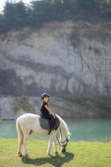 A person riding a white horse on a grassy field with a lake and cliffs in the background. 3