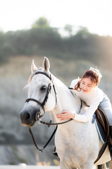 A person riding a white horse with a saddle, outdoors with a blurred natural background. 1