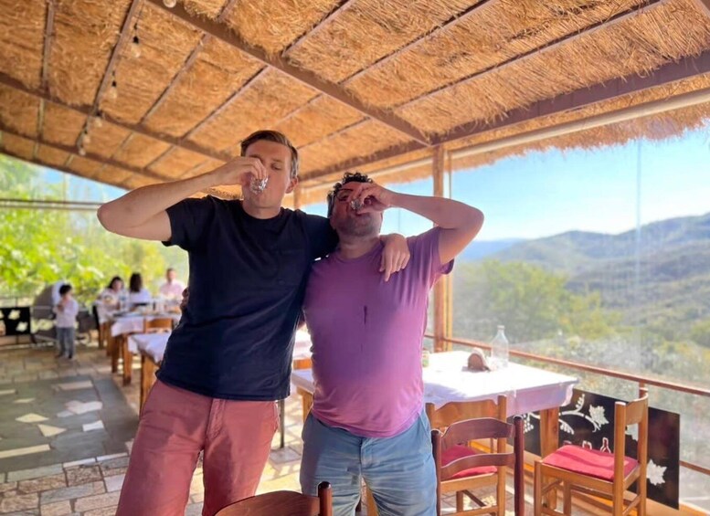 Two people standing under a thatched roof, with a mountainous landscape in the background. 4