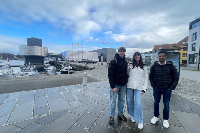 Three people standing on a paved area with a marina and modern buildings in the background. 5