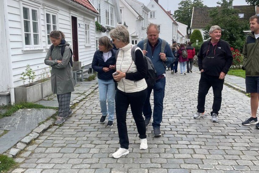 A cobblestone street with people walking and standing, white houses with red roofs, and a small tree in the foreground. 2