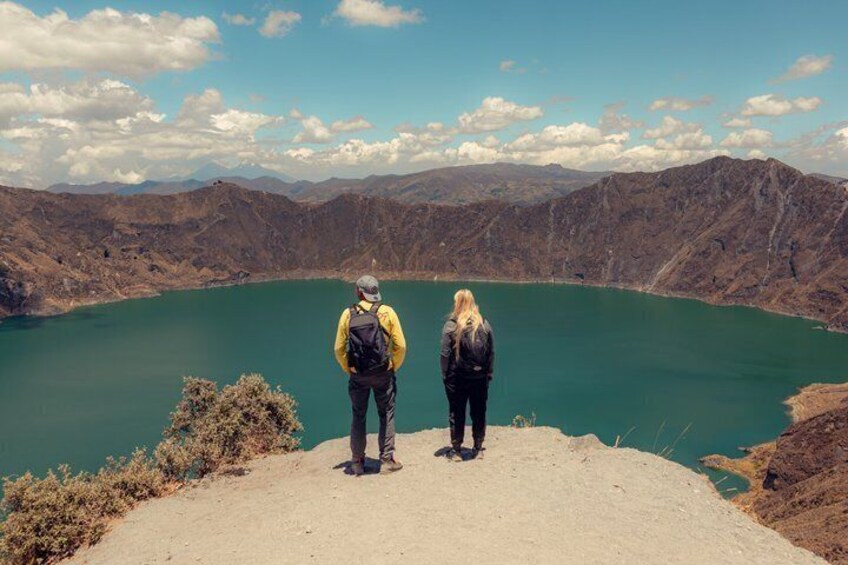 Two people standing on a hill overlooking a large body of water surrounded by mountains. 2