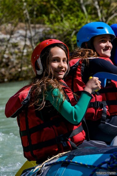 Two people in red life jackets on a blue raft, wearing helmets, on a river. 4