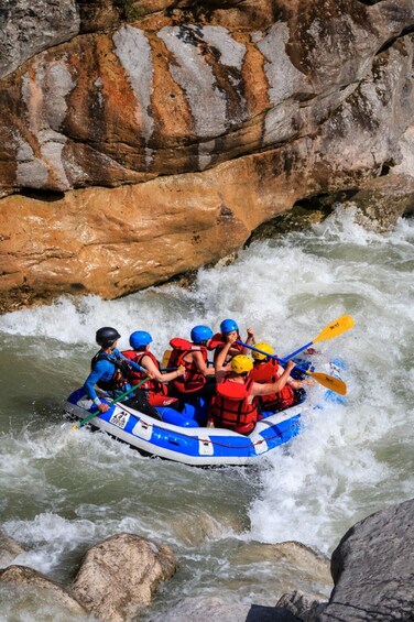 A group of people in a blue and white raft navigating through a river with white water rapids, surrounded by rocky terrain. 3