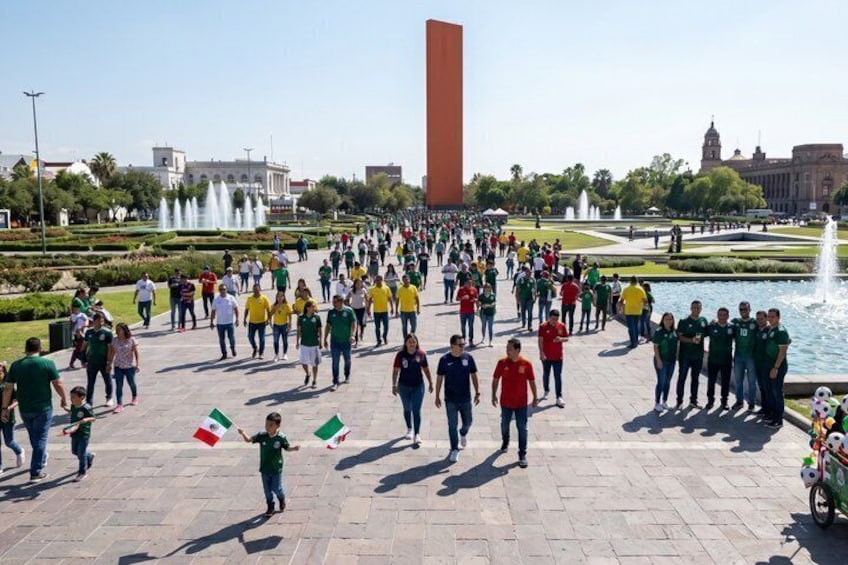 A large group of people in a plaza with fountains and a tall red monument. 2