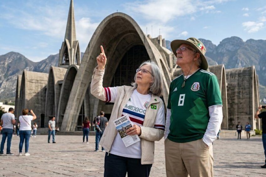 Two people in front of a large, arched building with mountains in the background. 1