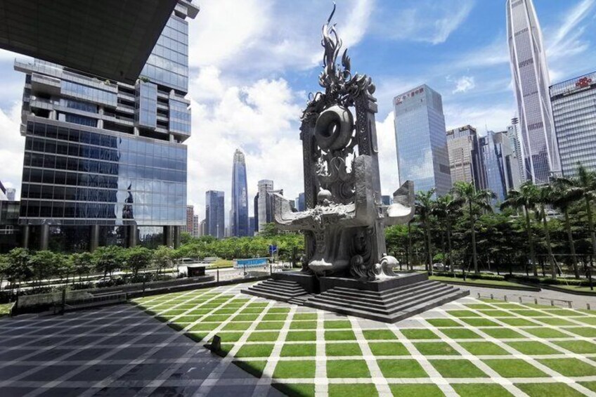 A city square with a central sculpture, surrounded by modern buildings and a clear sky. 1