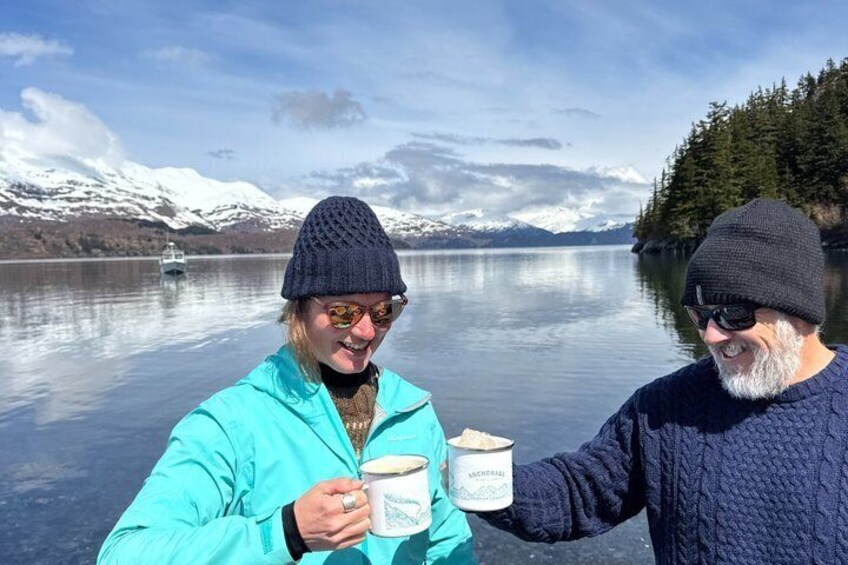 Two people holding mugs by a calm lake with snow-capped mountains in the background. 5
