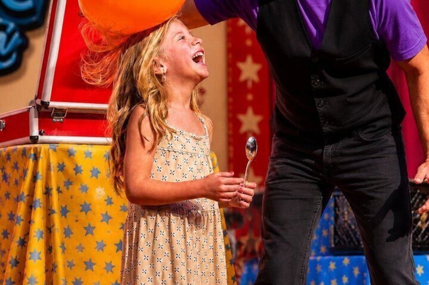 A child holding a spoon, a red suitcase, and a balloon on a stage with a star-patterned tablecloth. 3
