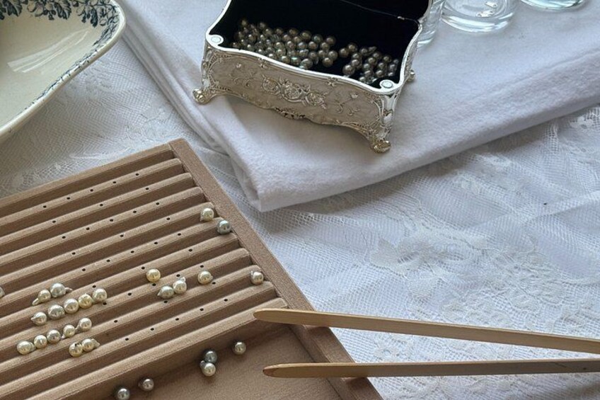 A wooden pegboard with beads, a small ornate box with beads, and a pair of wooden tongs on a white cloth. 1