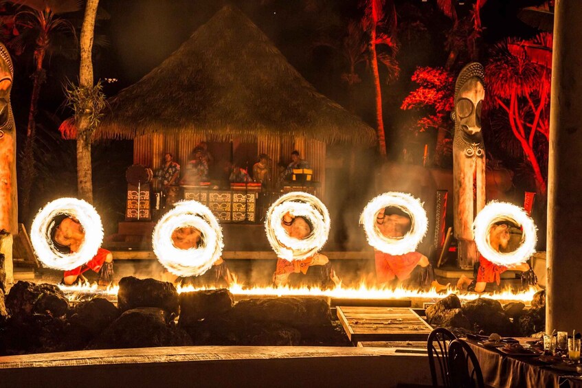 A fire performance with performers in traditional attire, a thatched-roof hut, and a tiki statue in a tropical setting. 1