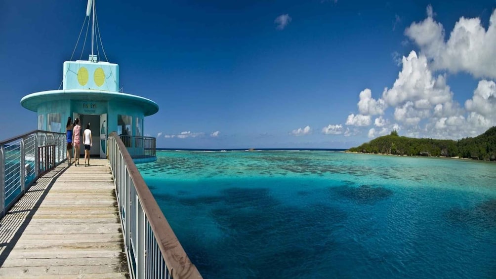 A pier leading to a turquoise building with a circular observation deck, surrounded by clear blue waters and a distant island. 3