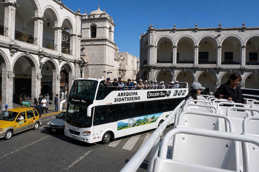 A double-decker bus labeled 'AREQUIPA SIGHTSEEING' and 'Cruzero Bus' parked in front of a historic building with arches. 1