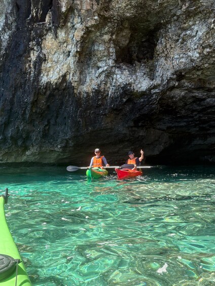 Two people kayaking in clear water with a rocky cave in the background. 3