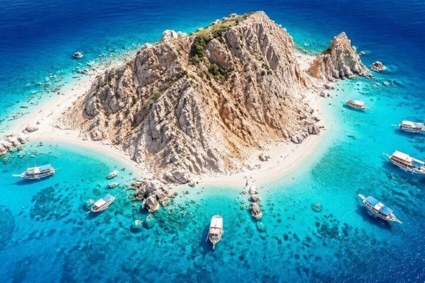 A rocky island surrounded by turquoise waters with several boats anchored near the shore. 1