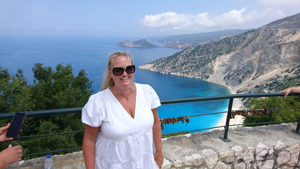 A person standing on a stone platform overlooking a coastal landscape with a clear blue sea and distant islands. 3