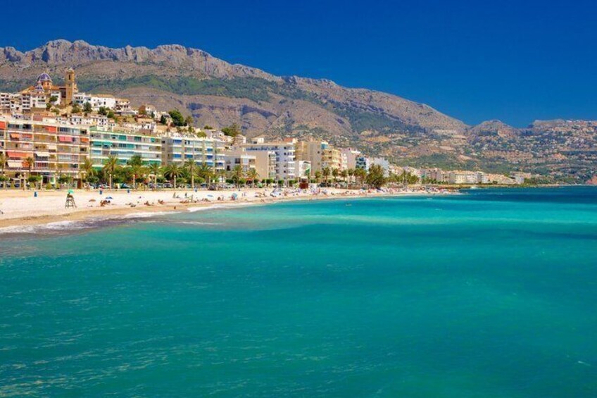 A coastal town with a beach, clear turquoise waters, and mountains in the background. 3