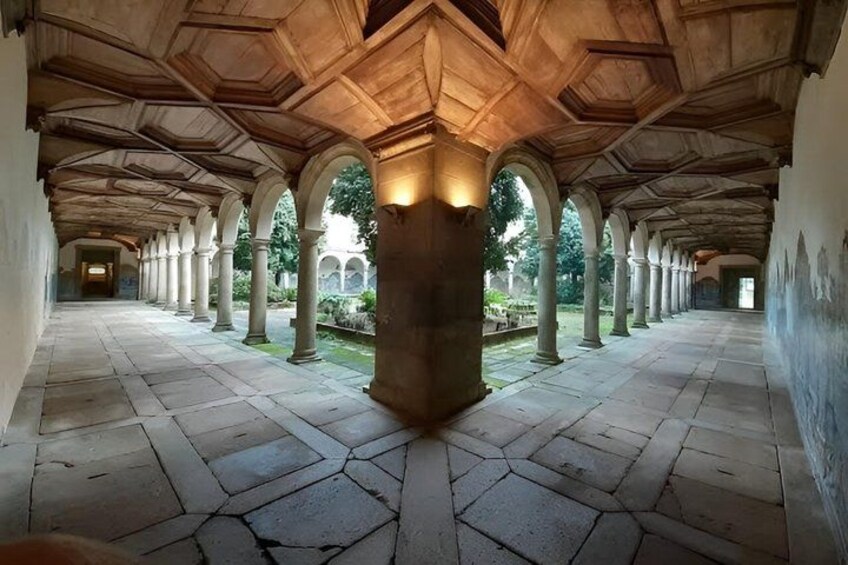 A covered corridor with stone flooring and wooden ceiling beams, featuring a central column and arches. 1