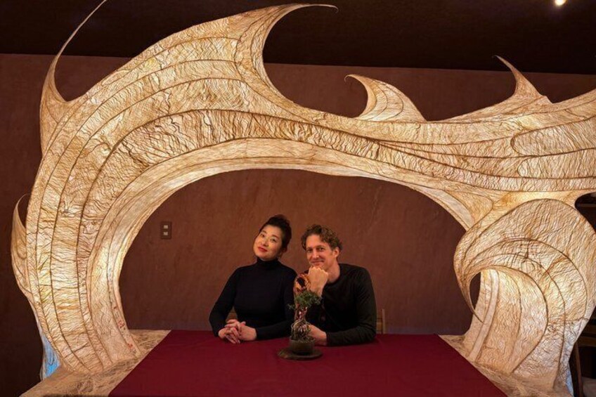 Two people seated at a table with a large, ornate, light-colored sculpture behind them. 1