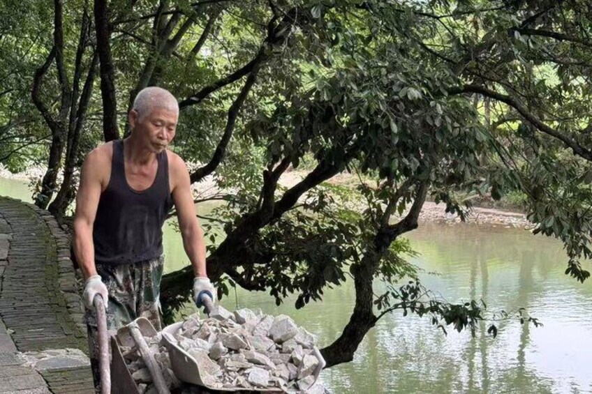A person pushing a wheelbarrow filled with rocks near a body of water with trees in the background. 4