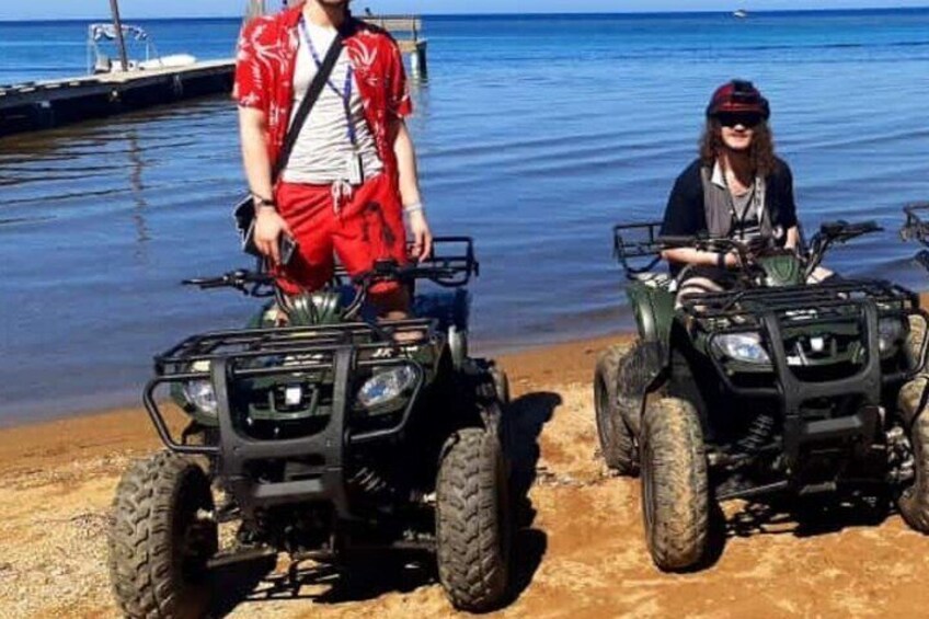 Two people on ATVs by a calm beach with a dock in the background. 5