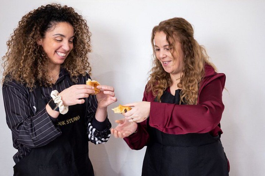 Two people wearing aprons, one with curly hair and the other with straight hair, holding pieces of food. 3