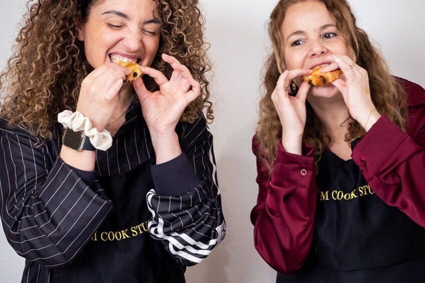 Two individuals wearing 'M Cook Studio' branded tops, one with curly hair and the other with straight hair, both holding a pizza slice. 2