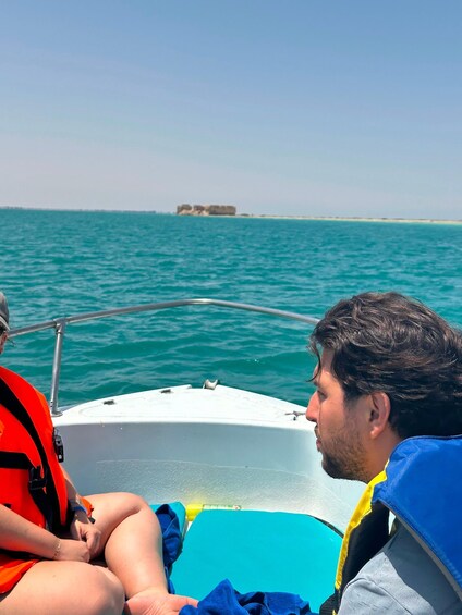 A boat with two people wearing life jackets, one in orange and the other in blue, on a turquoise sea with a distant island. 2