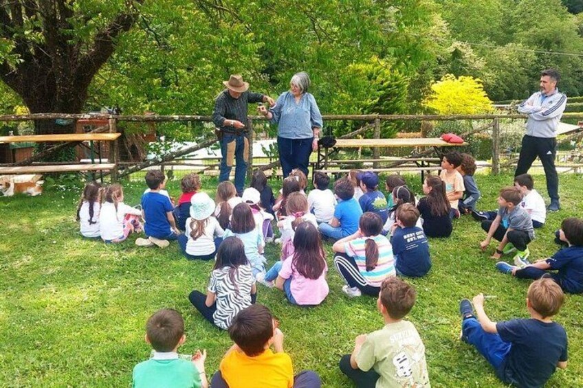A group of children sitting on grass outdoors, facing an adult who appears to be leading a group activity. 2