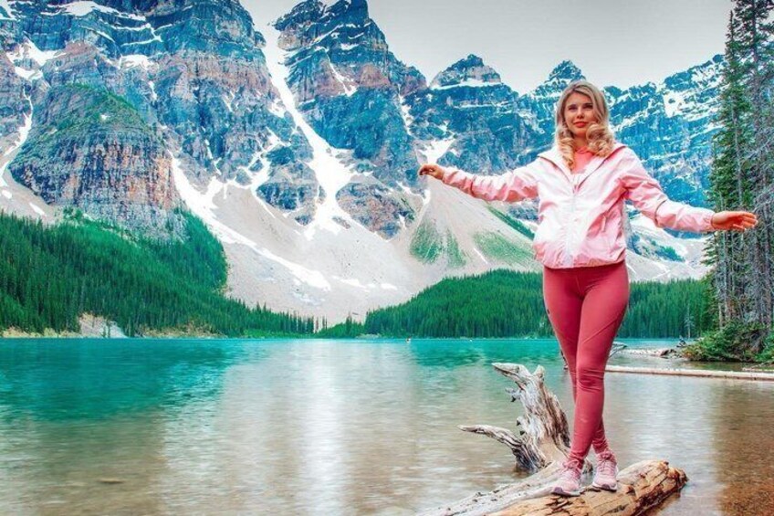 A person standing on a log in a lake with mountains in the background. 3