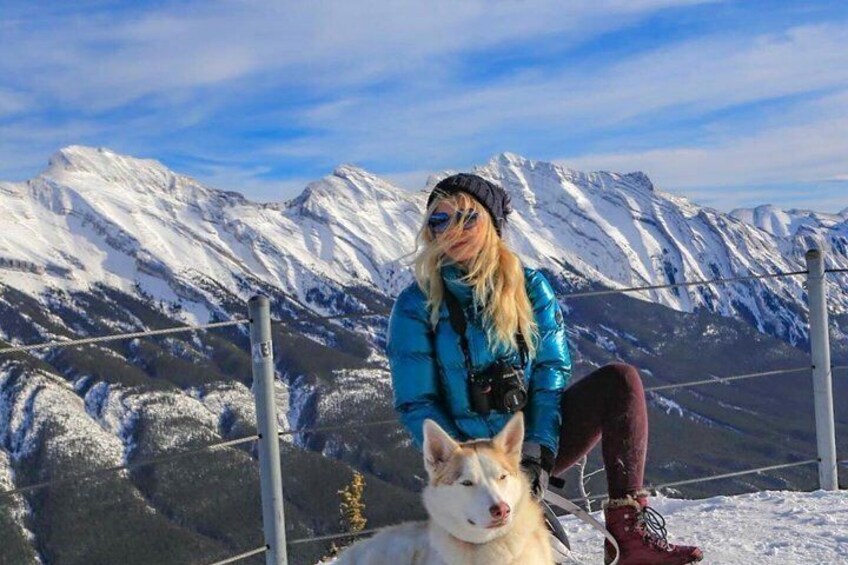 A person in a blue jacket and red boots sitting on a snowy ledge with a dog, overlooking snow-covered mountains. 2
