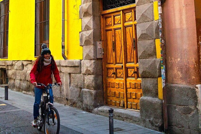 A person riding a bicycle on a cobblestone street, with a yellow building and a wooden door in the background. 4