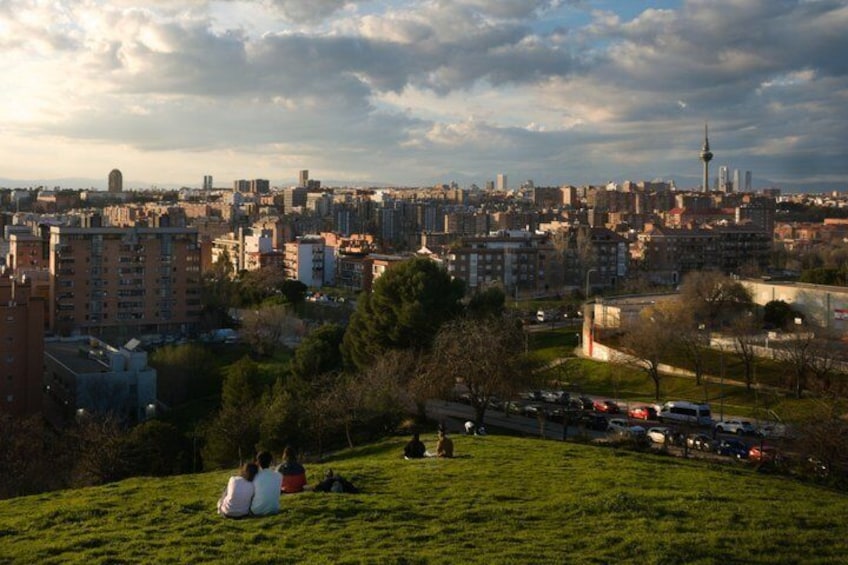 A cityscape with a prominent tower, a grassy hill with people, and a parking area. 3