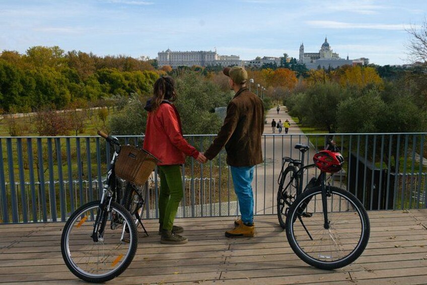 Two people holding hands on a wooden bridge with bicycles, overlooking a park and buildings in the background. 5