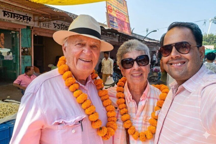 Three people wearing pink shirts and orange garlands, standing in front of a shop with a sign in a foreign language. 1
