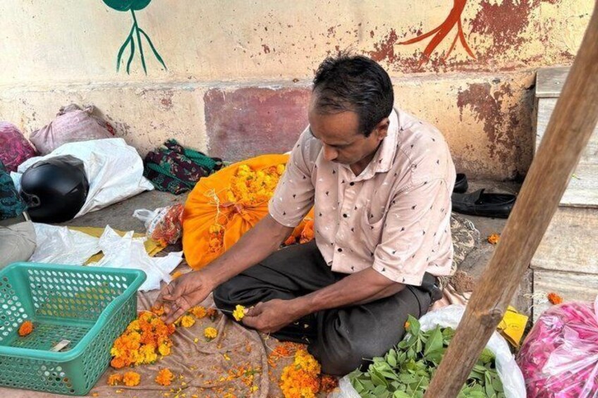A person sitting on the ground, surrounded by various items including a green basket, marigold flowers, and a plastic bag. 3