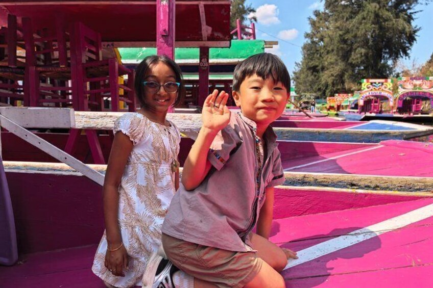 Two children on a colorful playground with a pink and blue floor, a wooden structure, and a green building in the background. 2