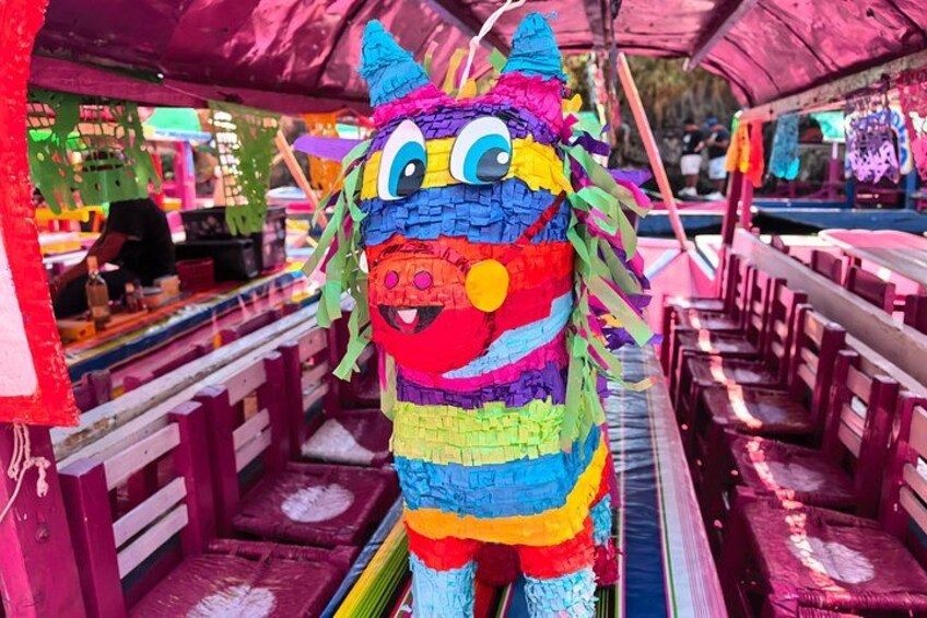 A colorful piñata with a face, hanging in an outdoor setting with wooden benches and a covered area. 2